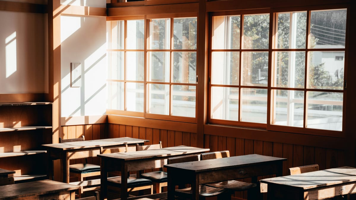 An empty wooden classroom with rows of desks and sunlight coming through tall windows